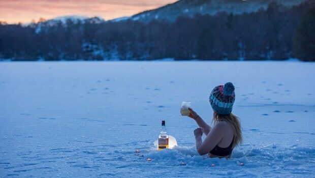 A thin white woman wearing a bikini top and a knit beanie stands chest-deep in a partially frozen lake, raising a glass. A bottle of alcohol is next to her, and the sun is setting behind mountains in the background.