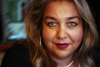 Close-up of a plump white woman with dark red lipstick, black eyeliner, wavy brownish-blonde hair, a pearl necklace, and hoop earrings