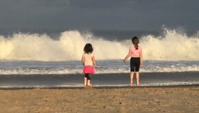 Two small children stand on the beach facing the ocean