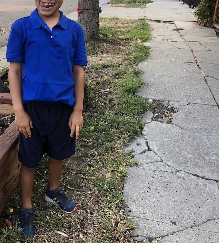 Photo of a 6-year-old boy from the chin down. He is making a goofy expression and wears a royal blue polo shirt with navy blue shorts. He stands next to a cracked sidewalk.