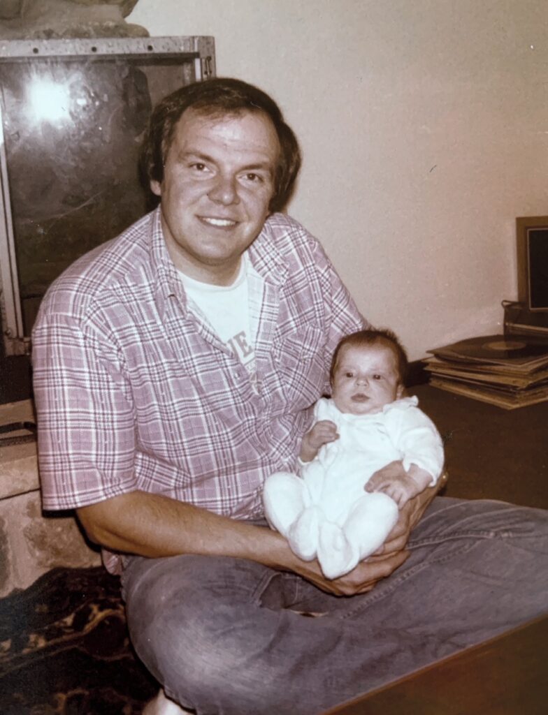 Photo circa the early 1980s, featuring Jennifer Acker's father, a white man in a plaid shirt, smiling as he holds his infant child