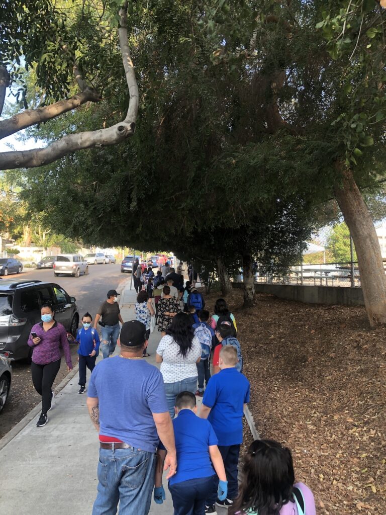 Parents and elementary school children wait in line beneath large trees outside a school campus