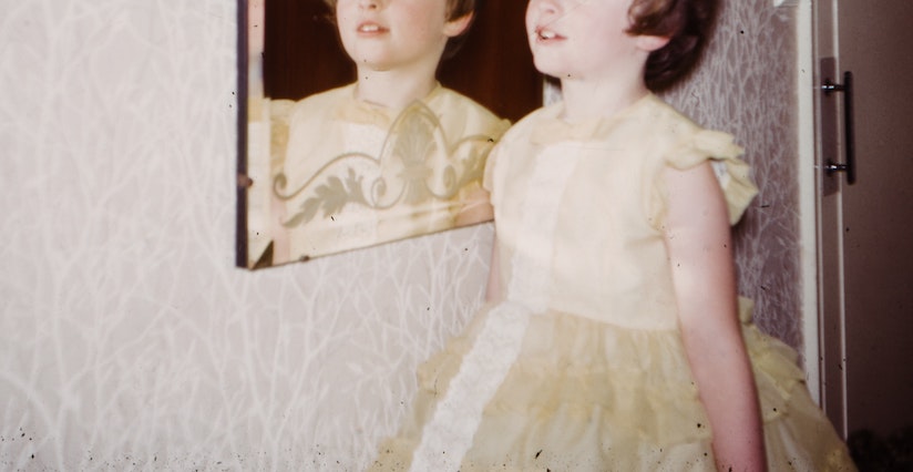 Young white girl with short brown hair and a frilly yellow dress standing next to a mirror