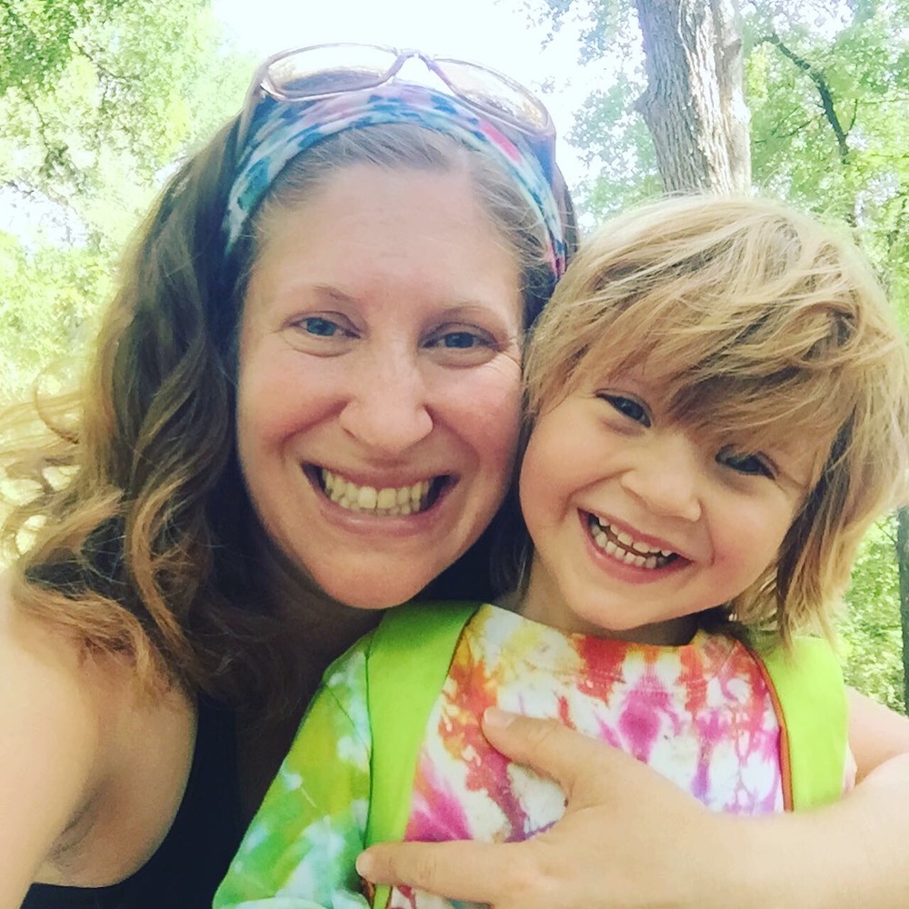 Smiling white woman with her arm around her young son. She wears a headband with glasses atop her head. He wears a tie-dyed shirt and has shaggy blond hair.