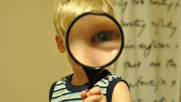 A blond-haired child of about 3 holds a magnifying glass in front of their face so all that's visible is one giant blue eye