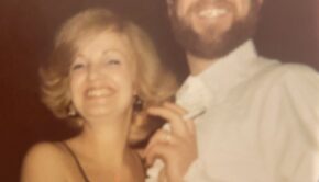 Close-up of a blonde-haired woman, circa the 1980s, with a big smile. She holds onto a bearded man with a cigarette.