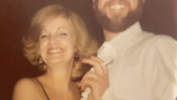 Close-up of a blonde-haired woman, circa the 1980s, with a big smile. She holds onto a bearded man with a cigarette.