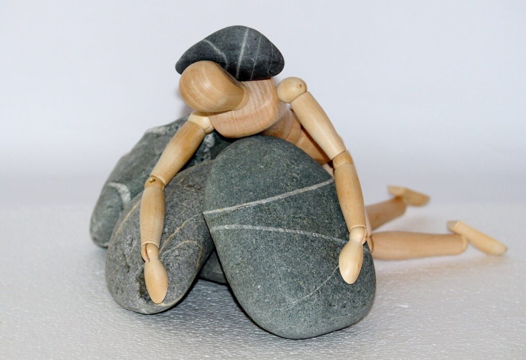 A wooden figure—the kind that artists use as models to draw people—leans over several smooth gray stones with white stripes encircling them. Another striped stone rests on the figure's back.