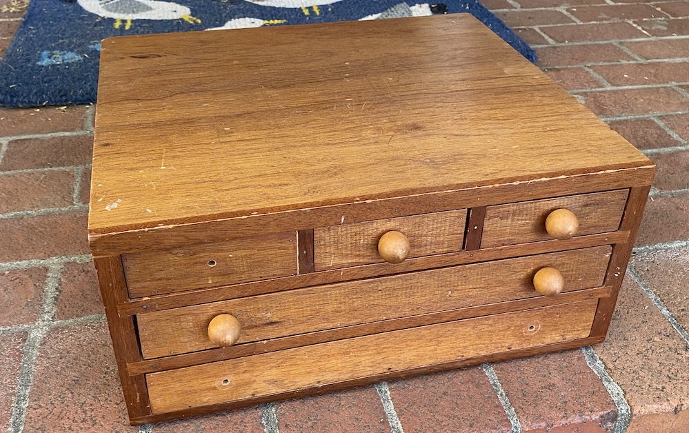 A small wooden chest of five drawers; three have round knobs, and the knobs are missing from two drawers