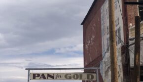 Side of an old brick building against a blue sky with white clouds. A worn sign extending from the building says "Pan for Gold."