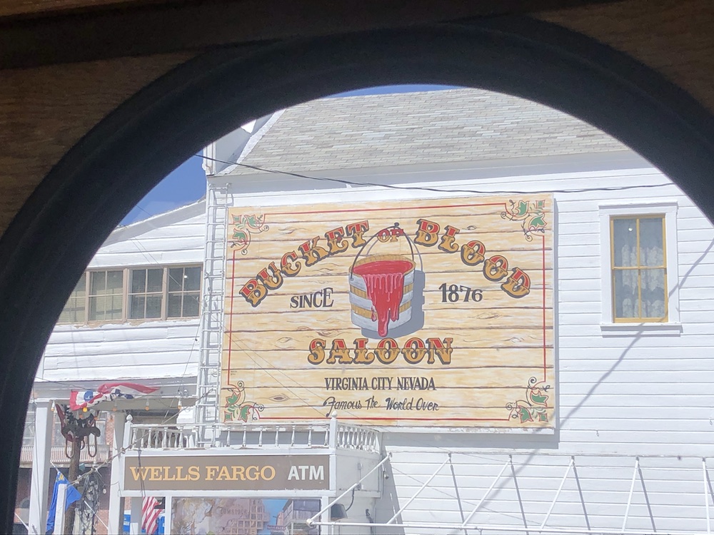 Through a rounded trolley window, a sign that says "Bucket of Blood Saloon: since 1876, Virginia City, Nevada" is visible on a wood-sided white building