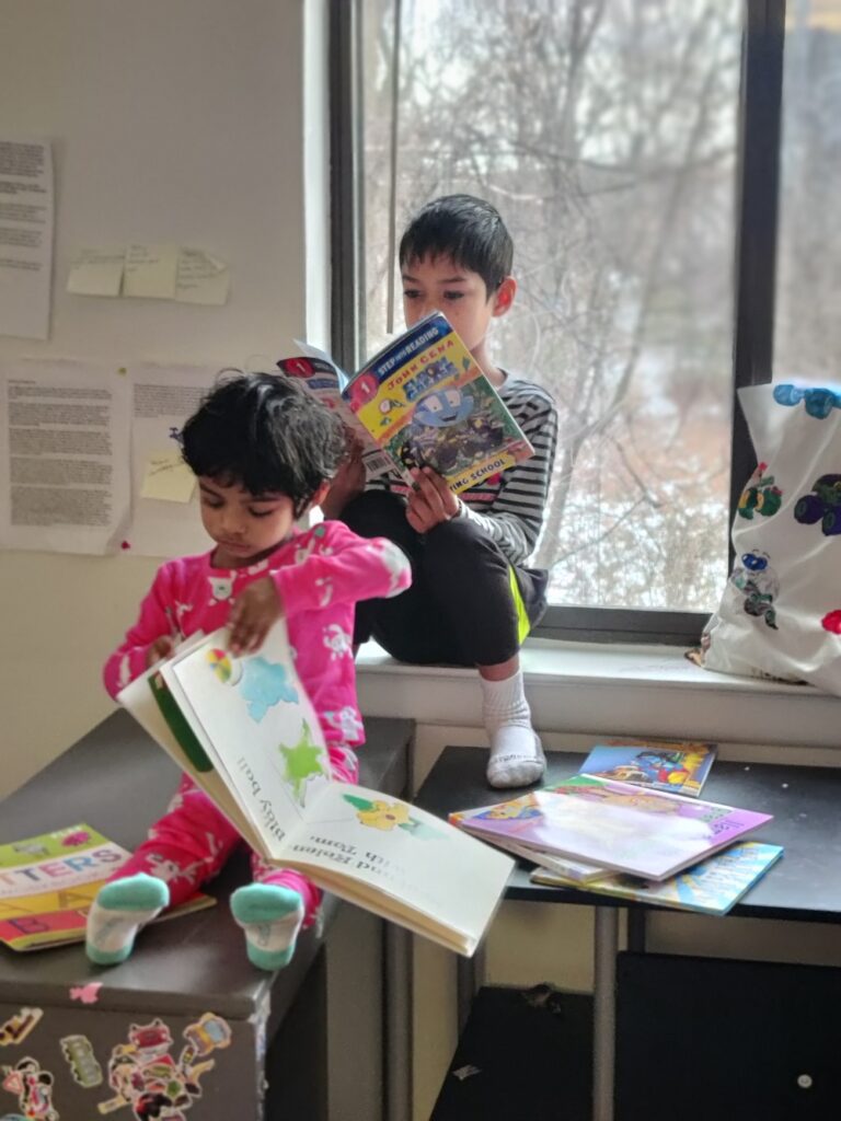 A preschool-aged child with medium brown skin and messy black hair in pink pajamas sits on top a metal desk, reading a picture book. The child's older sibling—lighter skinned with short, dark hair—sits behind them reading an early-readers book.