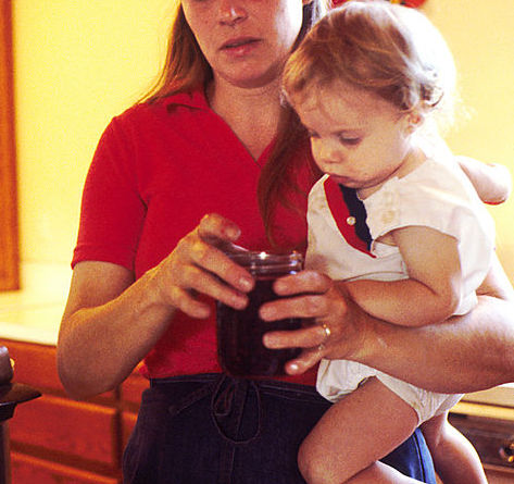 A mother with light skin, straight blonde hair, and a red polo shirt holds a blonde-haired toddler and a jar of jam