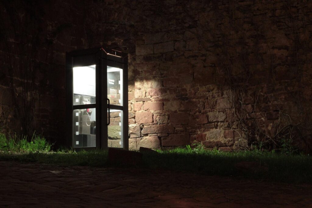 A phone booth lights up an otherwise dark landscape, partially illuminating a stone wall