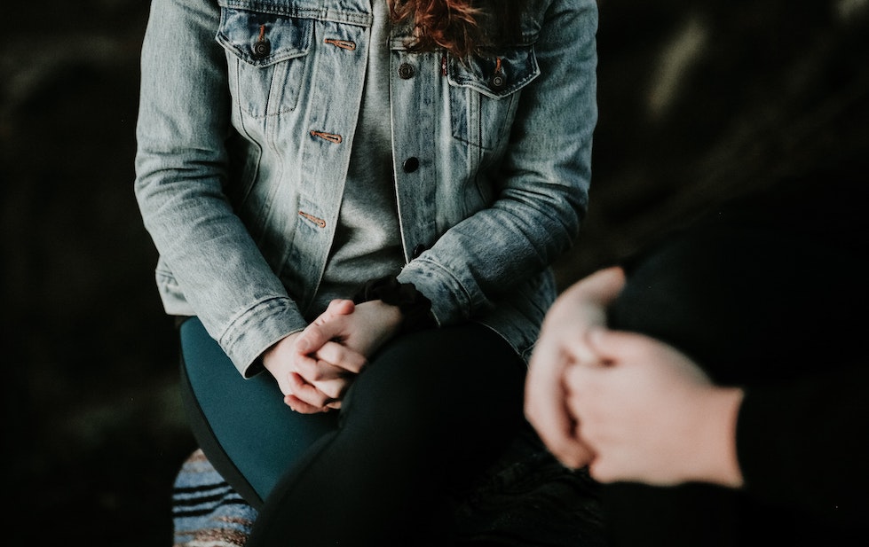 Photo depicting a light-skinned woman from the torso downward, her hands clasped. She wears a denim jacket and dark teal pants. To the right, another set of clasped hands can be seen.