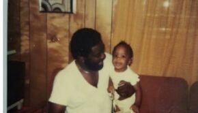 Vintage photo of a dark-skinned man in a white V-neck T-shirt looking lovingly at a smiling toddler he's holding. The toddler has medium-brown skin and short black curls and a ruffled white outfit.