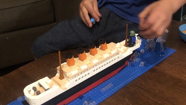 A child's light brown hands hover over a LEGO model of the Titanic