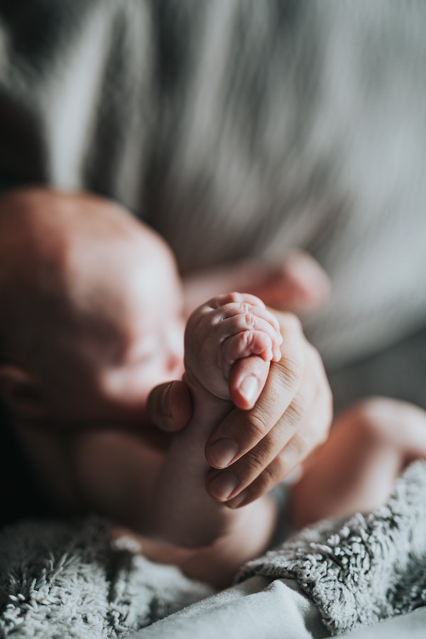 Close-up of a baby's hand holding an adult's hand; the baby can be seen in soft focus in the background