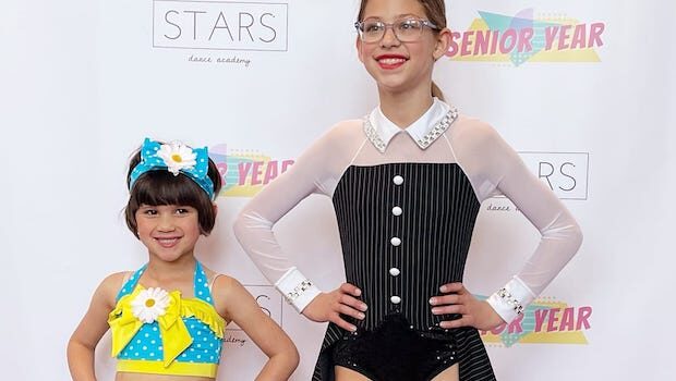 Two girls in dance uniforms. The younger girl has dark bobbed hair and a blue and yellow top and matching hat. The older girl has glasses, a low ponytail, and a black and white outfit.