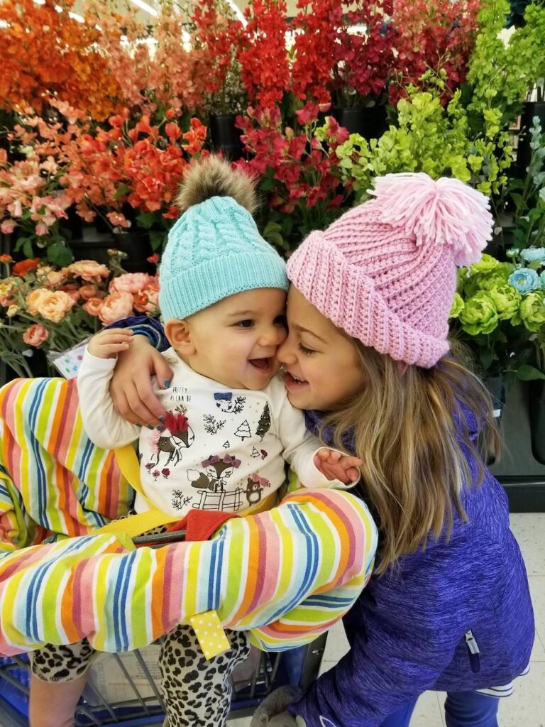 Young girl in a pink knit cap hugs a baby in a blue knit cap against a backdrop of autumn leaves
