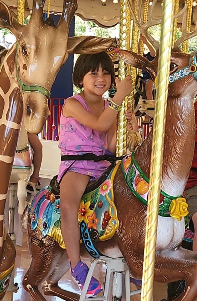 A girl of about five years old, with light brown skin and a dark brown bob, sits on a carousel animal
