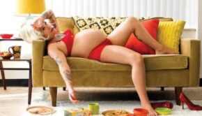 A pregnant woman in red lingerie lays on a yellow couch surrounded by plates and cups