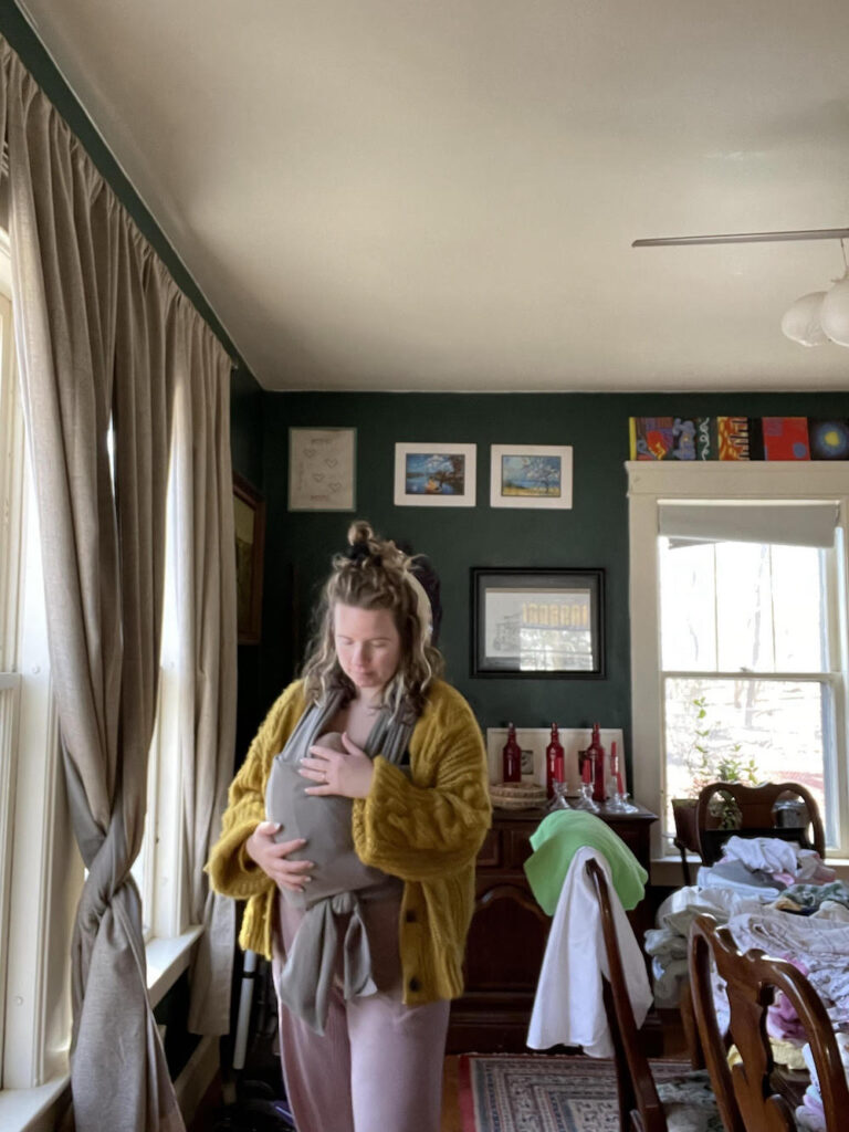 A woman wearing a yellow sweater holds a baby in a gray wrap-around carrier