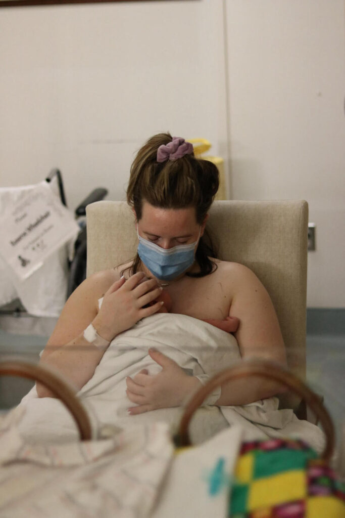 A mother with light skin and reddish brown hair holds a baby to her bare skin behind an incubator