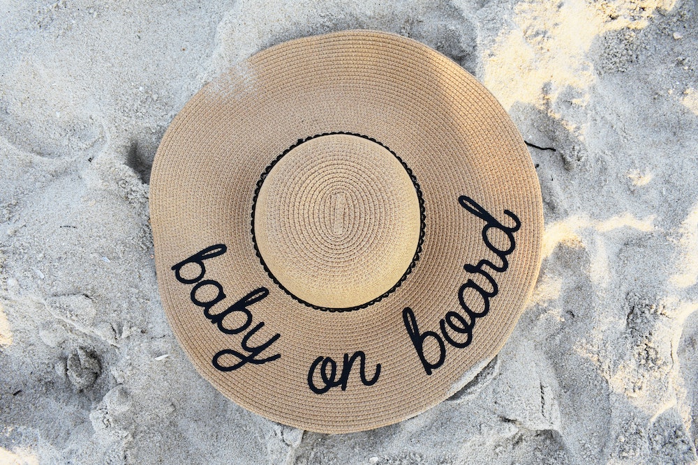 Straw hat with the words "baby on board" embroidered in black sits on the sand