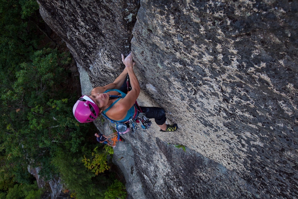 A woman with a focused expression on her face, blue tank top, pink helmet, and climbing harness, scales a vertical rock face. A man and green trees can be seen behind her.