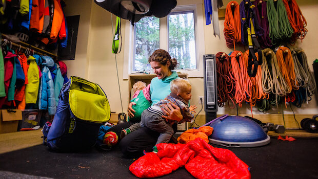 A mother holding toddler twins in eaach arm kneels on the floor of a room outfitted with climbing gear