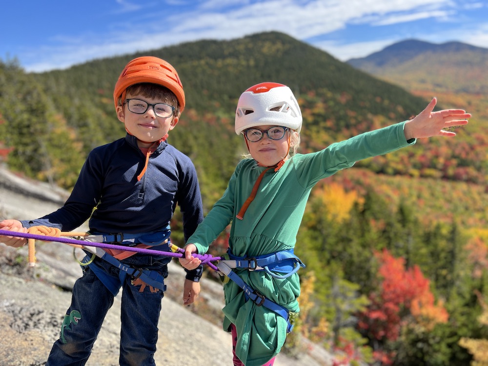 A boy and a girl, approximately 4 years old, in rock climbing gear against a background of mountains covered in fall foliage