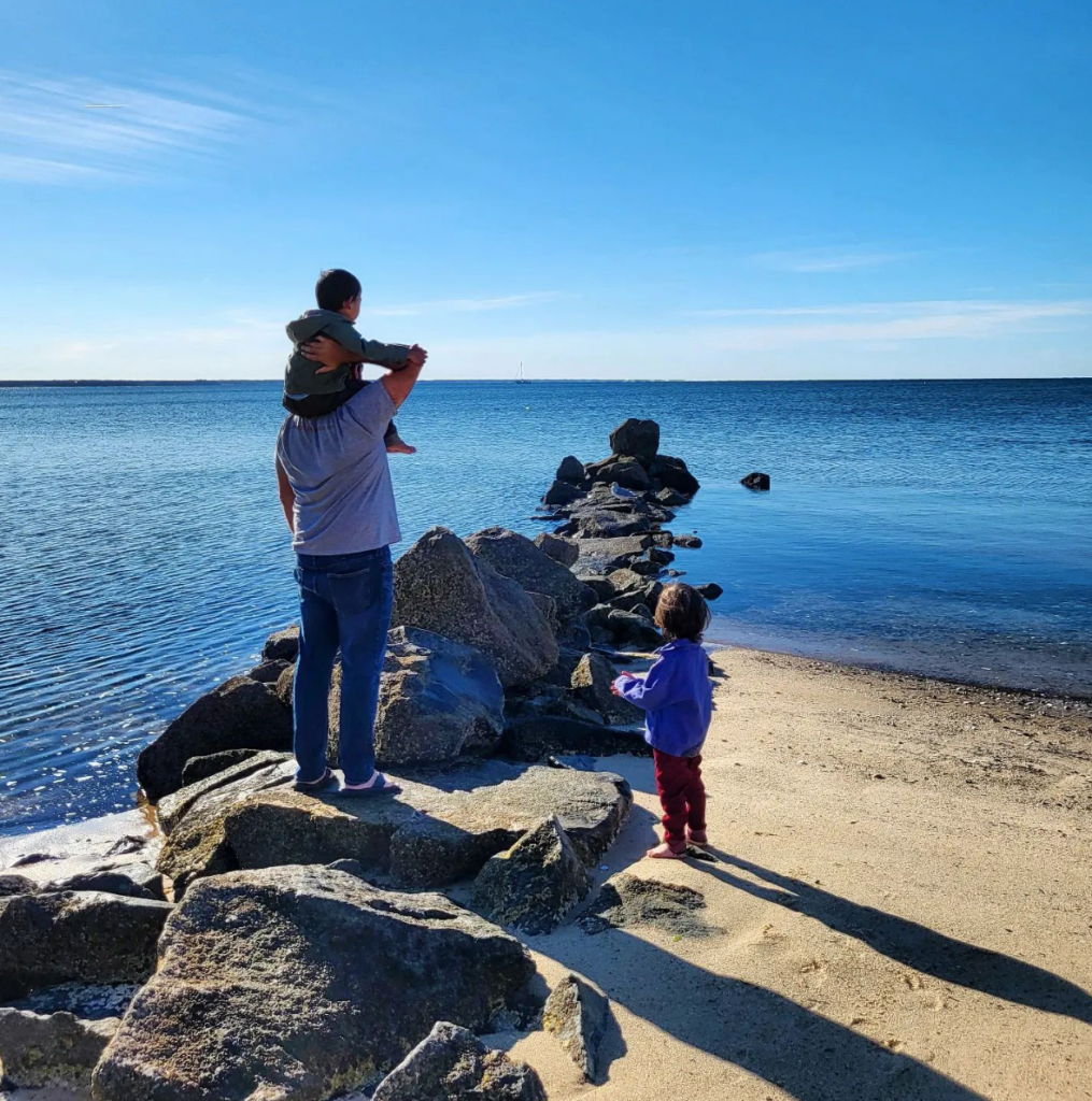 A father and two small boys stand on a rocky beach and face a large body of water
