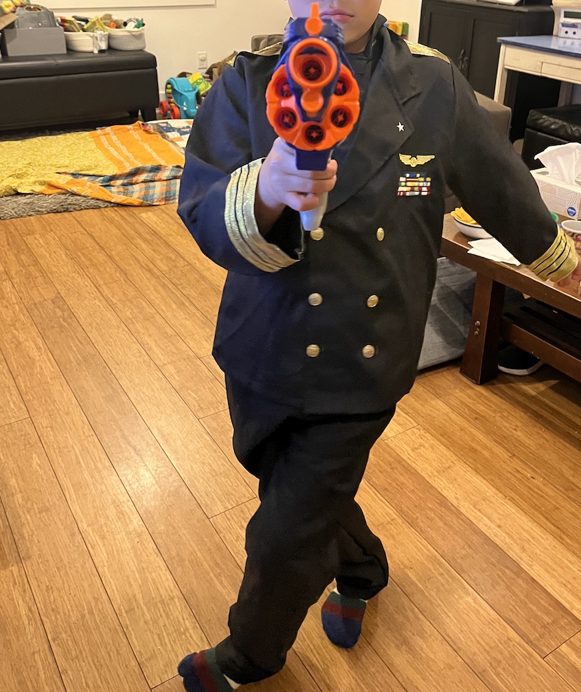 An eight-year-old boy dressed in a naval officer's uniform points a Nerf gun at the camera