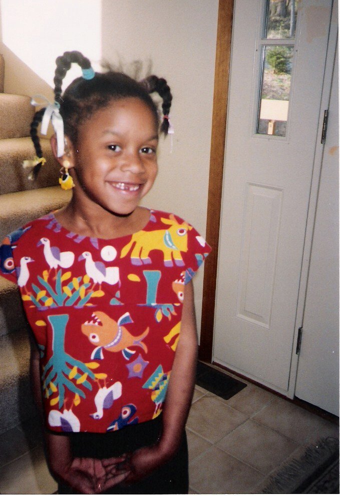 Five-year-old Angela wears a red shirt with fanciful animals on it and smiles, showing a missing tooth. Her hair is in braids with white ribbons.