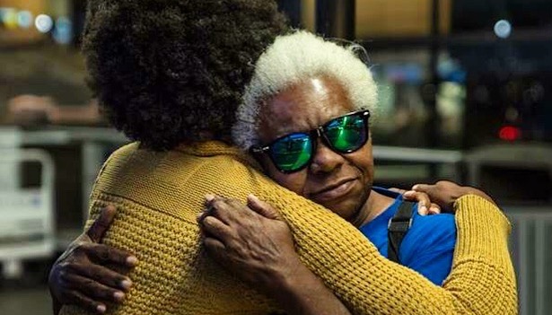 A younger woman with a yellow sweater and an Afro hugs her mother, a woman with white hair and sunglasses