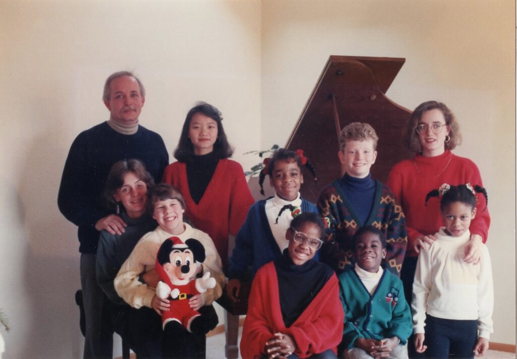 1980s family photo that depicts white parents and eight children of multiple races, standing in front of a piano
