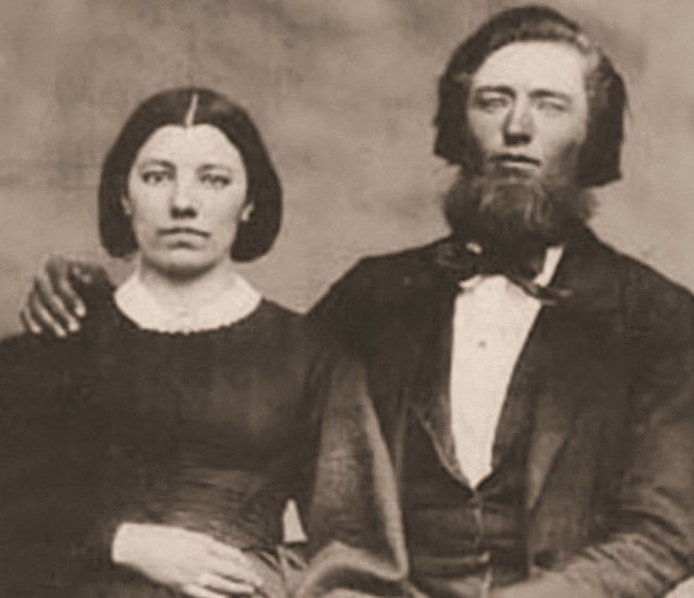 Caroline and Charles Ingalls. Caroline wears a dark dress with a white lace collar. Charles has a beard, longish hair, and a suit and bowtie. His hand rests on Caroline's shoulder.