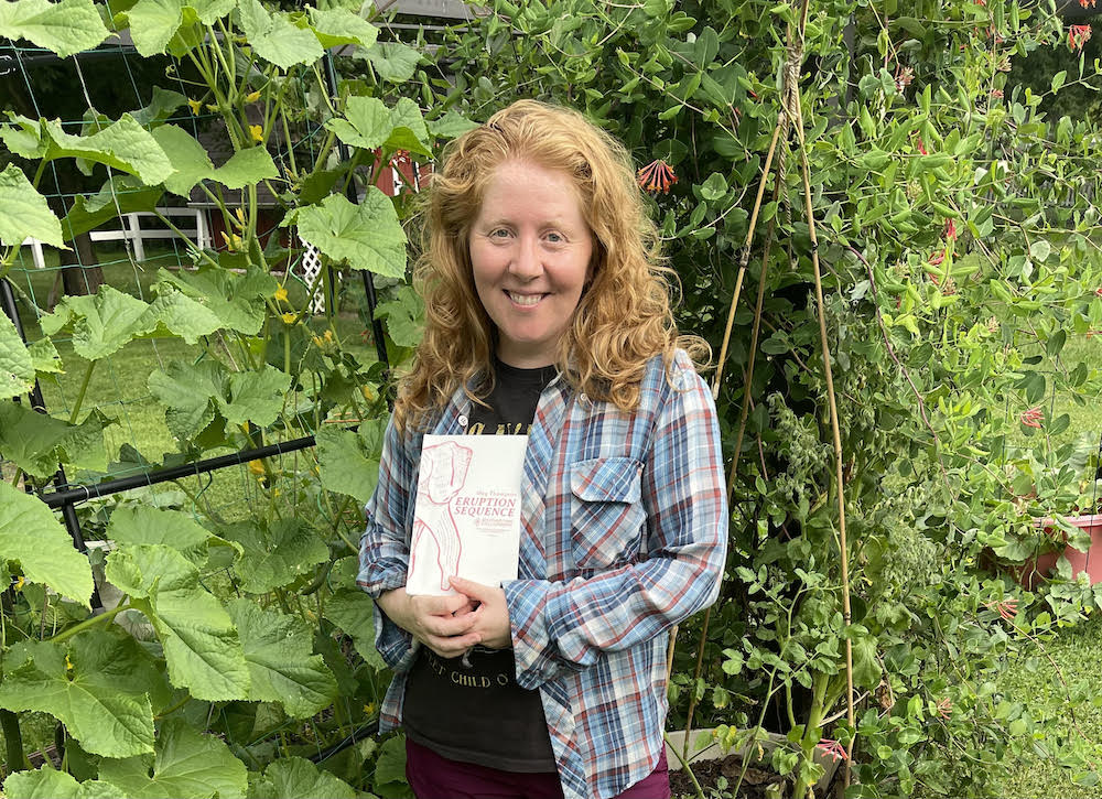 Author Meg Thompson stands in front of bright green vines, holding her book, Eruption Sequence. She has red hair and wears a plaid shirt over a T-shirt.
