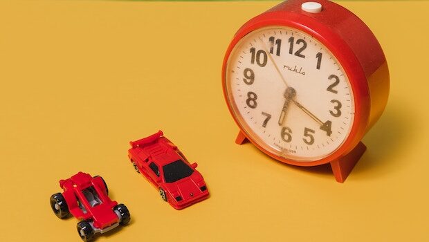 Red alarm clock showing 6:20 next to two small red cars on a yellow backdrop