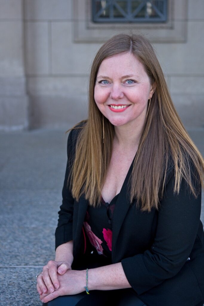 A light-skinned woman with straight blond hair and a black jacket sits with her hands folded