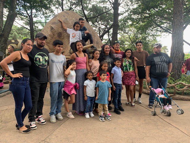 A family photo taken at a park in Guatemala. Eighteen people ranging from infancy to early middle age stand with their arms around each other. 