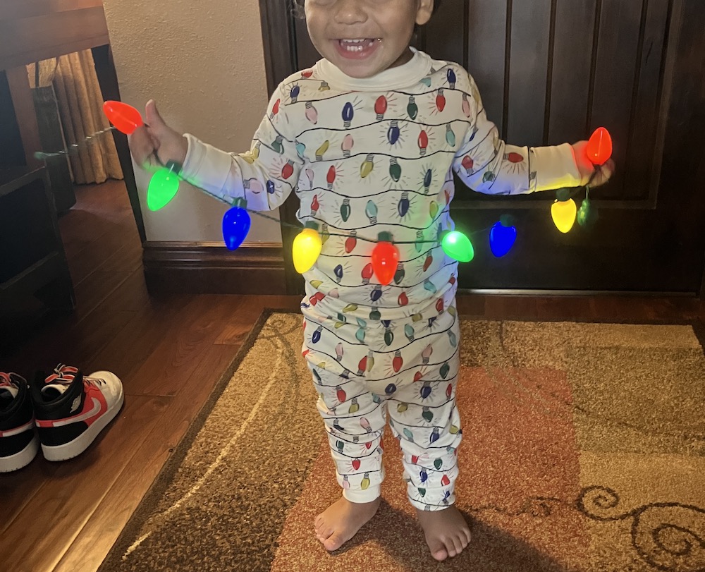 A toddler in Christmas light pajamas holds a string of Christmas lights