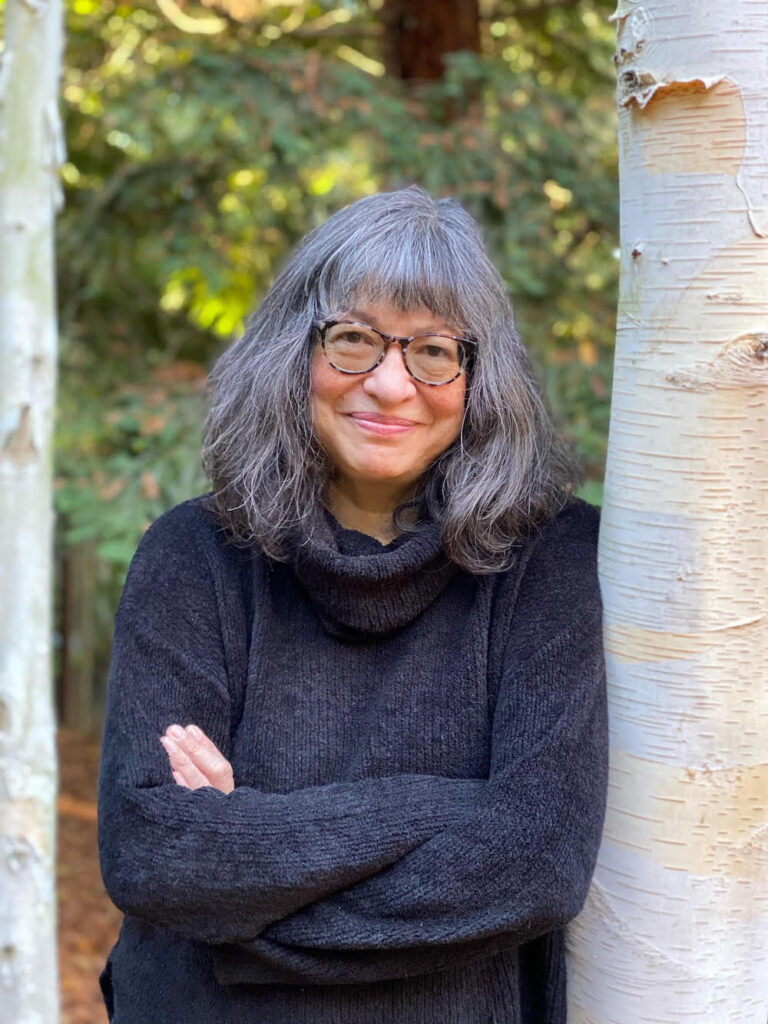 A biracial (Japanese and white) woman with gray shoulder length hair leans against a tree. She wears glasses, a black sweater, and a slight smile.