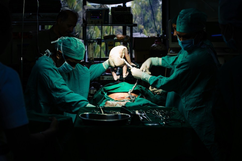 Medical staff in green scrubs lift a baby from a patient's abdomen in an operating room