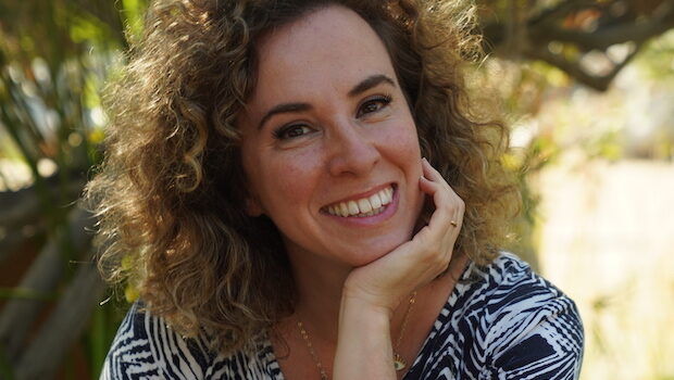 Author Katya Apekina sits outdoors wearing a black and white dress. She has light skin, curly brown hair, and is smiling while resting her chin in her hand.