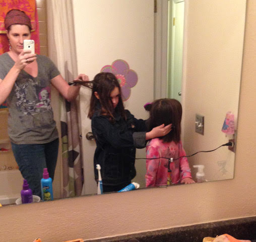 Mom in bathroom with two daughters. She is styling the oldest's hair as the oldest styles the youngest's.