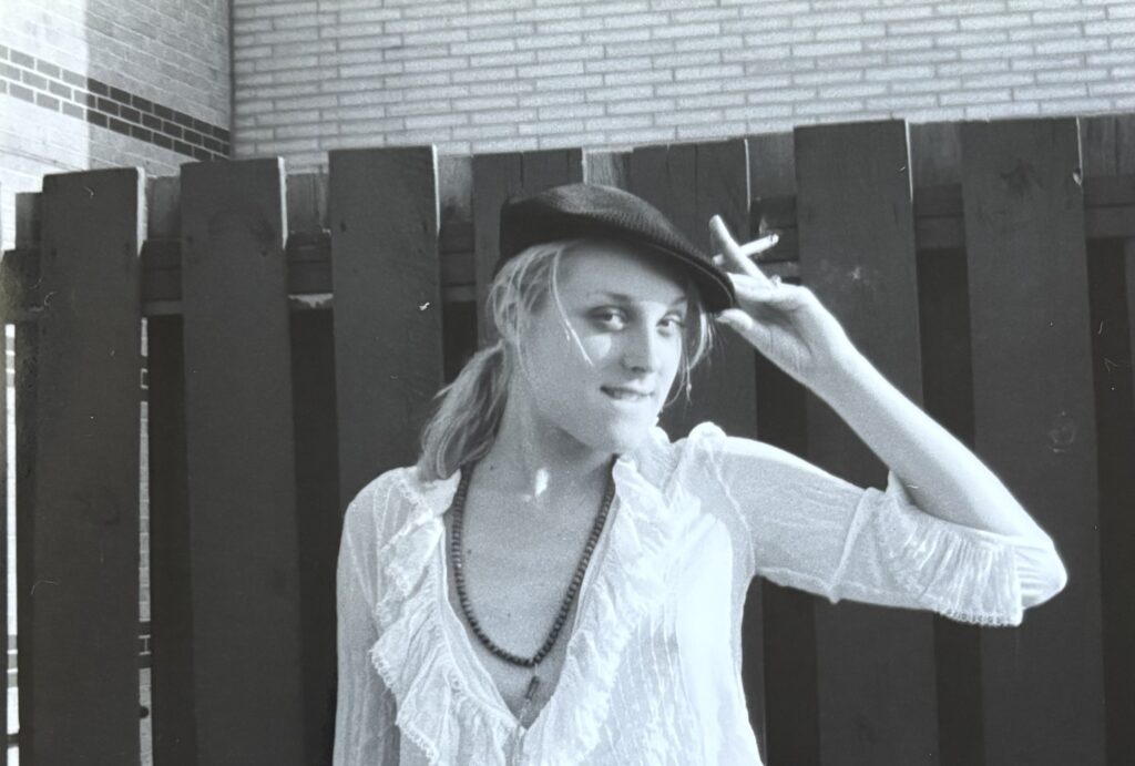 Young woman with a beret stands in front of a fence. She touches her hat with a hand that also holds a cigarette.