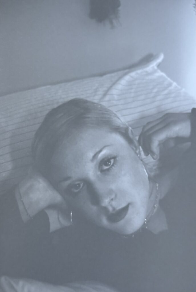 Close-up of a young woman with dark lipstick, long-sleeved shirt, and necklaces, hair pulled back, lying on a bed