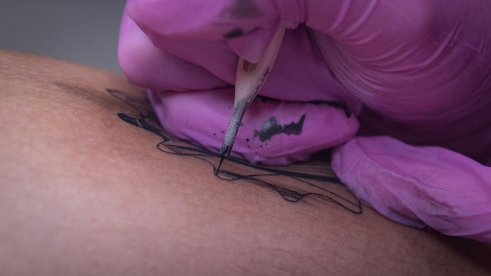 Hand wearing pink glove inking a gear-shaped tattoo on medium-light skin